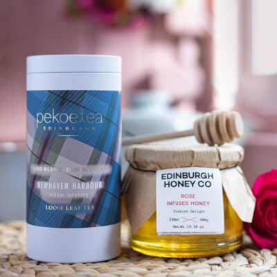 A Pekoetea tin labelled 'Newhaven Harbour herbal infusion loose leaf tea' with the text 'hand blended in scotland' next to a 400g jar of Edinburgh Honey Co. Rose Infused Honey with a honey dipper resting on top of the jar