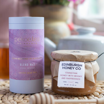 A Pekoetea tin labelled 'Blend No 3 herbal infusion loose leaf tea' with the text 'hand blended in scotland' next to a 400g jar of Edinburgh Honey Co. Cranachan Honey with Whisky and Raspberries with a honey dipper resting in front of the jar